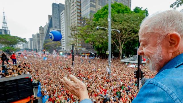 Lula, de perfil, sorri com os olhos fechados, vestindo camisa jeans azul, com a mão direita estendida para uma multidão de apoiadores em uma avenida urbana, com prédios altos e um balão azul ao fundo